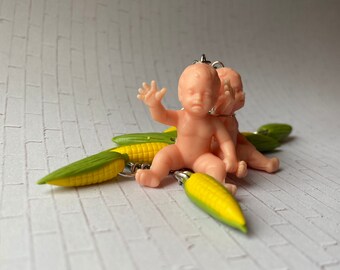 May include: A group of small, plastic baby figurines in a light peach color, sitting on top of several corn on the cob charms. The corn charms have yellow kernels and green husks, with silver-colored metal loops.