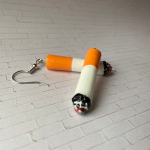 May include: Cigarette-shaped earrings with orange, white, black, and red details. The earrings have a silver fish hook style ear wire. The earrings are made to look like a lit cigarette.