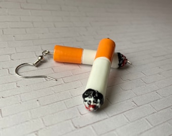 May include: Cigarette-shaped earrings with orange, white, black, and red details. The earrings have a silver fish hook style ear wire. The earrings are made to look like a lit cigarette.