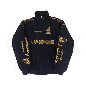 Puede incluir: Chaqueta negra con cremallera, costuras doradas y la palabra "Lamborghini" impresa en la parte delantera y las mangas. La chaqueta también presenta un logotipo dorado de Lamborghini con un toro en el pecho derecho.