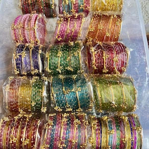 May include: A selection of colourful bangles in a clear plastic container. The bangles are in various colours, including red, pink, green, blue, and yellow. Each bangle is decorated with gold-coloured accents.