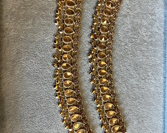 May include: Two gold-toned anklets with a teardrop and small bell design. The anklets have a chain closure and are displayed on a gray surface. The anklets are made of metal and have a shiny finish.