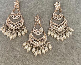 May include: Three gold-coloured chandelier earrings with a crescent moon design. Each earring features clear, sparkling crystals and a cluster of pearly white beads. The earrings are displayed on a grey surface.