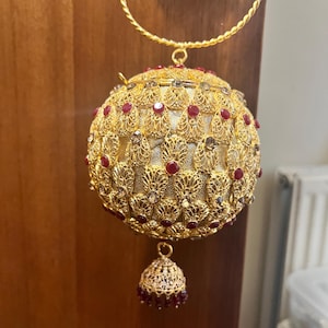 May include: A gold-coloured, orb-shaped handbag with intricate filigree and red gemstone accents. The bag has a matching handle and a dangling bell-shaped charm. The design is ornate and detailed, suggesting a formal or special occasion accessory.