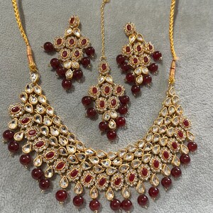 以下が含まれることがあります： ネックレス、イヤリング、ヘアアクセサリーがセットになったゴールドトーンのジュエリーセット。赤とクリアの宝石と赤いビーズのアクセントで飾られています。ネックレスとイヤリングは涙のしずくのデザインです。