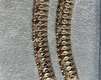 May include: Two gold-toned anklets with teardrop-shaped mirrored accents and small, round gold beads. The anklets also feature clear, sparkling embellishments and delicate chain extensions.