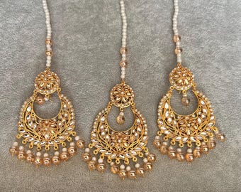 May include: Three gold-toned tikka headpieces with detailed floral and geometric patterns. Each piece has a crescent-shaped pendant decorated with clear crystals and dangling beads. The headpieces have a pearl-like chain.