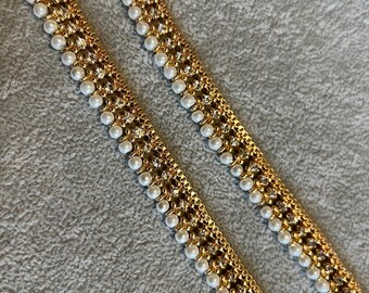 May include: Two gold-colored decorative trims with alternating rows of small white pearls and clear rhinestones. The trim has a repeating pattern and is set against a gray background. The trim is likely used for embellishing clothing or accessories.