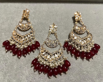 May include: Three gold-toned chandelier earrings with intricate detailing. Each earring features a crescent-shaped design adorned with clear crystals and small white pearls. The bottom of each earring is embellished with a cluster of deep red teardrop-shaped beads.