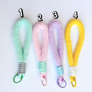 May include: Four fluffy wrist lanyards in pastel colors: mint green, pink, lavender, and yellow. Each lanyard has a coordinating clasp and a decorative band at the base. The lanyards are numbered 1 through 4.