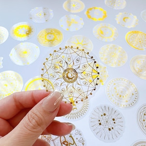 May include: A collection of circular transparent stickers featuring intricate gold foil designs of suns, moons, stars, and geometric mandalas. Each sticker displays detailed celestial patterns with thin lines and metallic finishes on a clear backing.
