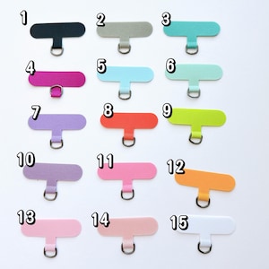 May include: A collection of fifteen colorful phone charm tabs in various shades, including black, gray, teal, pink, blue, green, orange, purple, and white. Each tab has a metal ring for attaching to a phone case.