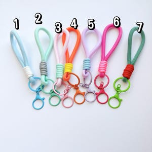 Può includere: Sette portachiavi in corda colorata con chiusure a moschettone in metallo, numerati da uno a sette. Ogni portachiavi presenta un cordino ad anello in azzurro, menta, rosa, arancione, lavanda, fucsia o verde, con componenti metallici e nodi abbinati.