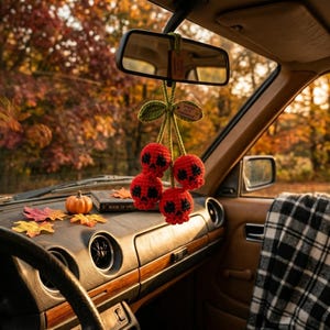 Könnte beinhalten: Ein gehäkeltes Auto-Accessoire am Rückspiegel, bestehend aus drei roten, schädelförmigen Kugeln mit schwarzen Augen- und Nasendetails, verbunden durch grüne Stiele und Blätter, mit einem kleinen Holzanhänger mit der Aufschrift SPROUT CUTE HANDMADE.