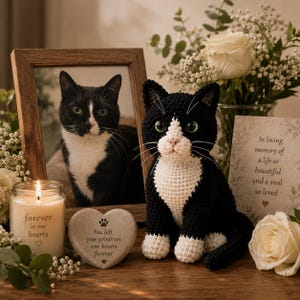 May include: A black and white crocheted cat figurine sits beside a wooden picture frame containing a photo of a black and white cat. A heart-shaped stone with the text 'You left paw prints on our hearts forever' and a candle labeled 'forever in our hearts' are placed nearby.