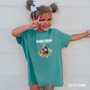 A young girl wearing a green t-shirt with a Mandalorian-themed design is smiling and making a peace sign with her fingers.