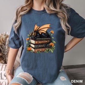 A woman wearing a blue t-shirt with a graphic design of a dragon sitting on a stack of books, surrounded by flowers.