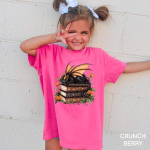 A young girl wearing a pink t-shirt with a dragon design and books on top, smiling and making a peace sign with her hand.