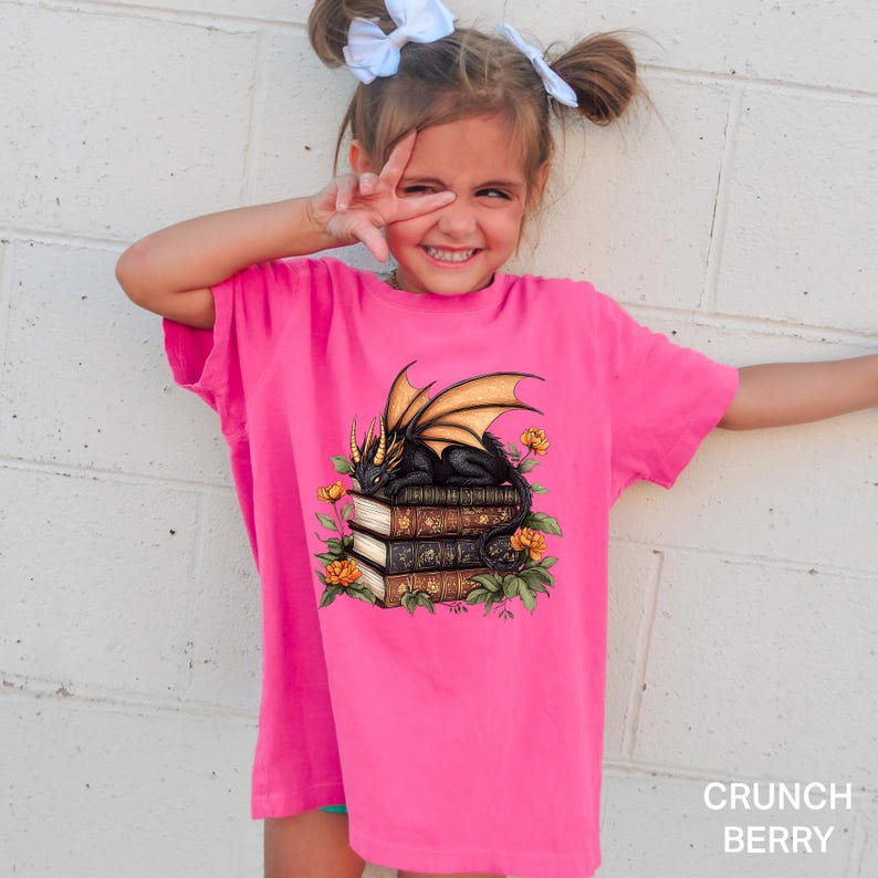 A young girl wearing a pink t-shirt with a dragon design and books on top, smiling and making a peace sign with her hand.