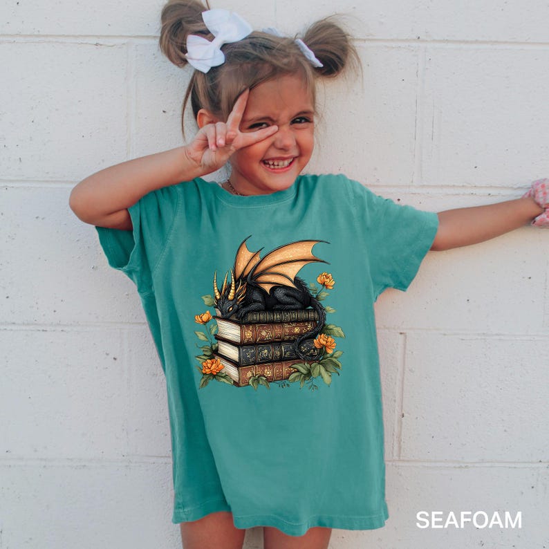 A young girl with pigtails is smiling and making a peace sign with her fingers, wearing a green t-shirt with a dragon and books design.