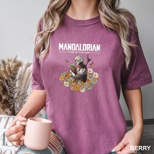A woman wearing a purple t-shirt with a graphic of a character from the Star Wars series, specifically Mandalorian, holding a cup of coffee.