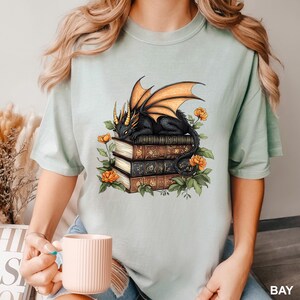 A woman wearing a t-shirt with a graphic design of a dragon perched on a stack of books, surrounded by flowers. She is holding a white mug in her hand.