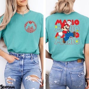 May include: A teal t-shirt featuring a small red circular graphic on the front chest composed of various Mario icons. The back displays a large graphic of Mario jumping with the text MARIO repeated in red outline font and yellow stars.