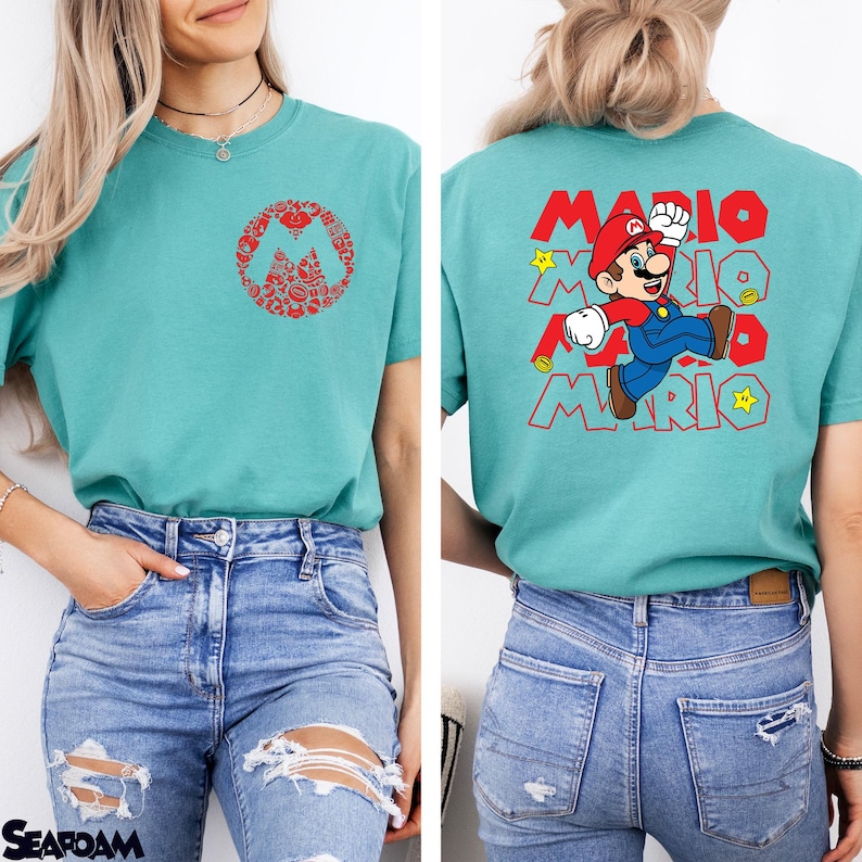 May include: A teal t-shirt featuring a small red circular graphic on the front chest composed of various Mario icons. The back displays a large graphic of Mario jumping with the text MARIO repeated in red outline font and yellow stars.
