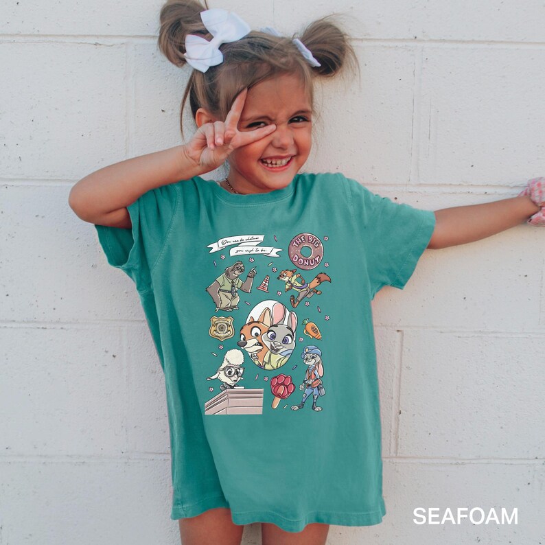 A young girl wearing a green t-shirt with a cartoon character design is smiling and making a peace sign with her fingers.