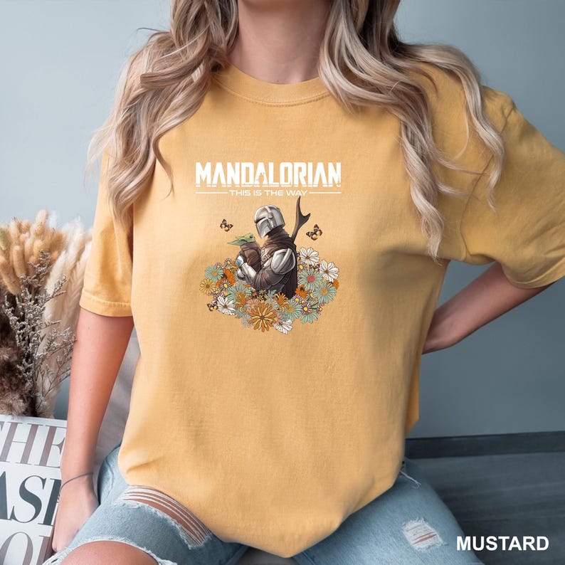 A person wearing a yellow t-shirt with a Mandalorian graphic and text printed on it.