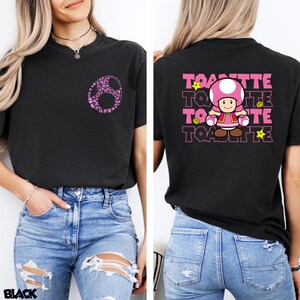 May include: A black t-shirt featuring a small pink circular graphic on the front left chest. The back displays a large graphic of the character Toadette, surrounded by yellow stars and coins, with the text Toadette repeated in pink block letters.
