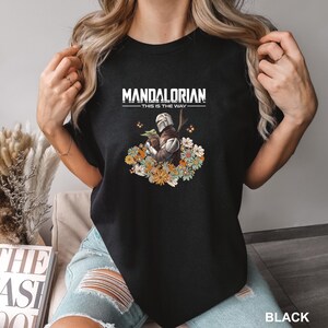 A woman wearing a black t-shirt with a graphic design of a character from the Star Wars franchise, specifically Mandalorian, surrounded by flowers.