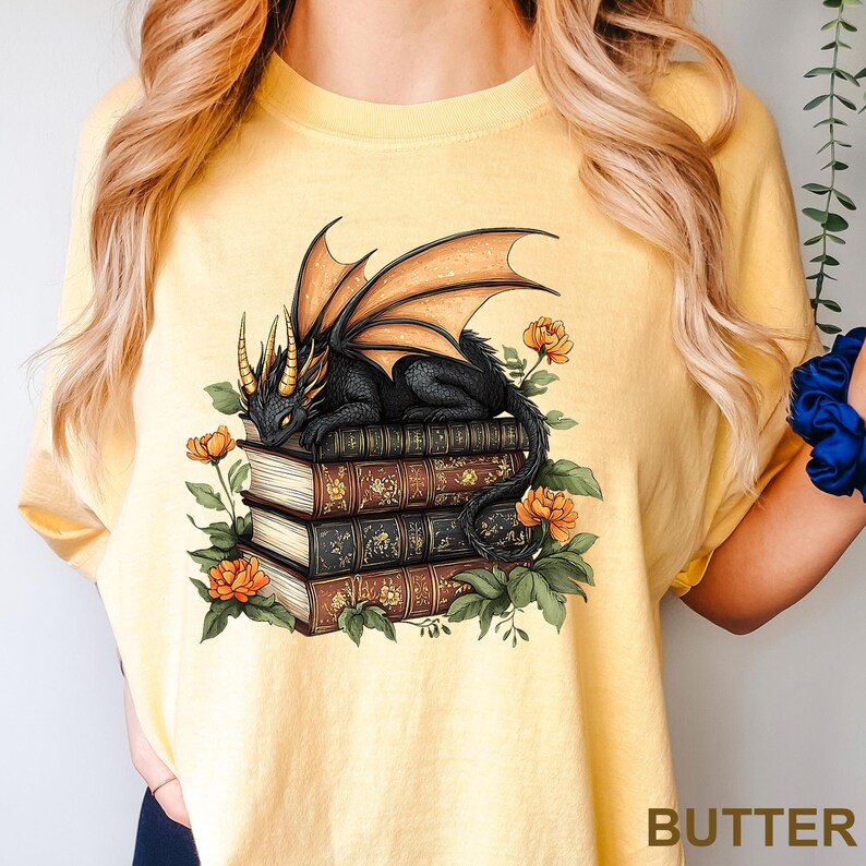 A yellow t-shirt with a graphic design depicting a black dragon perched on a stack of books, surrounded by flowers and leaves.