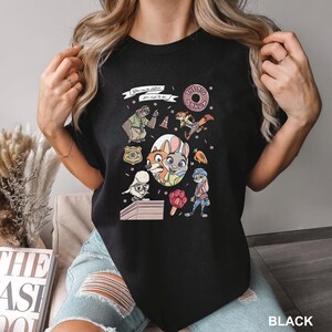 A woman wearing a black t-shirt with a colorful graphic design featuring cartoon characters and various objects.