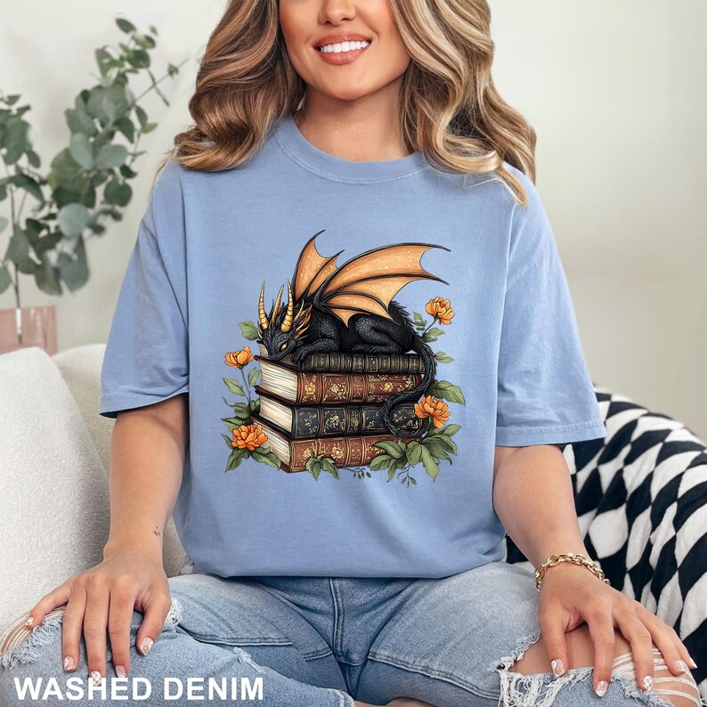 A woman wearing a blue t-shirt with a graphic design of a dragon perched on a stack of books, surrounded by flowers and leaves.