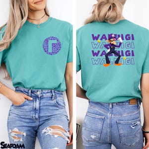 May include: A teal short-sleeve t-shirt featuring a purple circular graphic on the front chest containing small icons. The back displays a large purple graphic of the character Waluigi with the repeated text WALUIGI and small yellow stars.