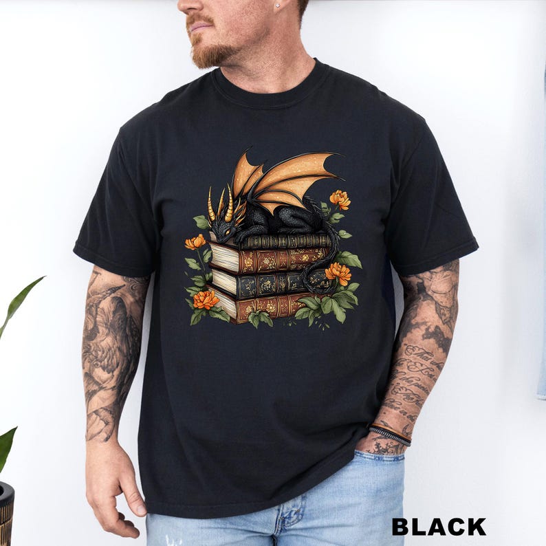 A man wearing a black t-shirt with a graphic design of a dragon perched on a stack of books, surrounded by flowers and leaves.