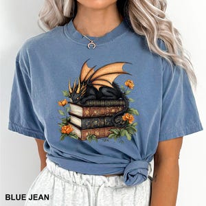 A woman wearing a blue t-shirt with a graphic design of a dragon holding a stack of books and surrounded by flowers.