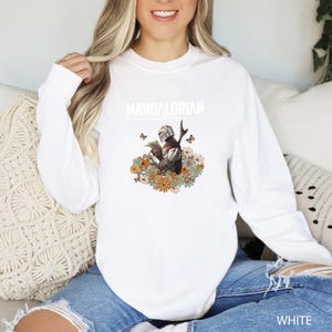 A woman wearing a white long-sleeved shirt with a graphic of a character from the Star Wars franchise, specifically the Mandalorian, sitting on a couch.