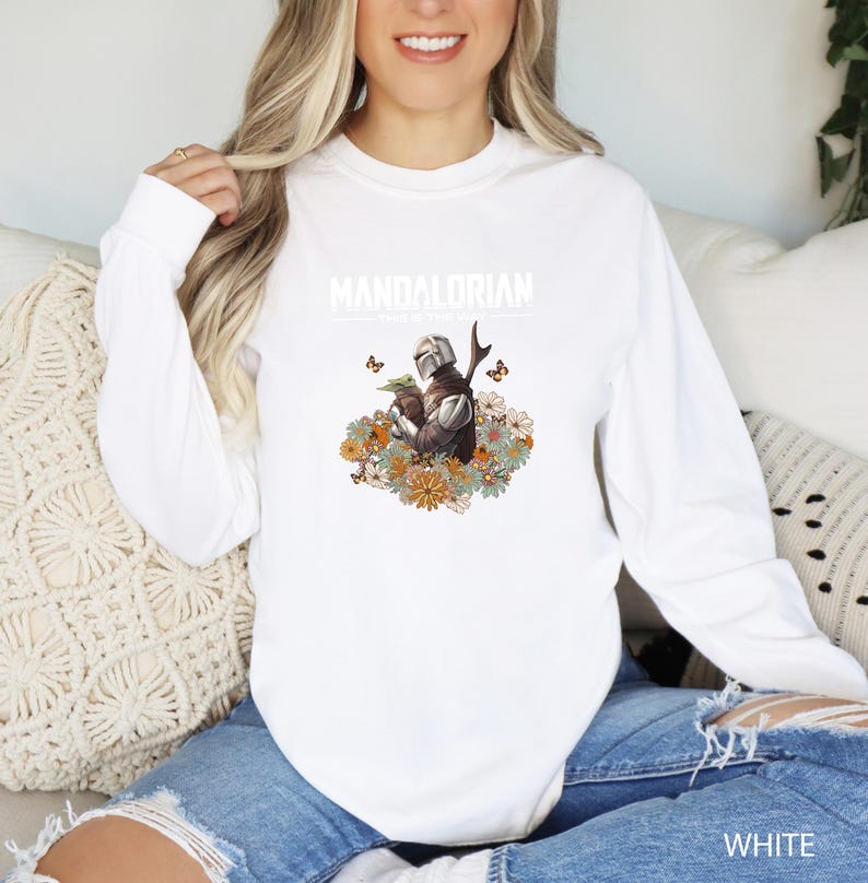 A woman wearing a white long-sleeved shirt with a graphic of a character from the Star Wars franchise, specifically the Mandalorian, sitting on a couch.