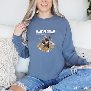 A woman wearing a blue sweatshirt with a graphic of a character from the Star Wars franchise, specifically Mandalorian, sitting on a couch.