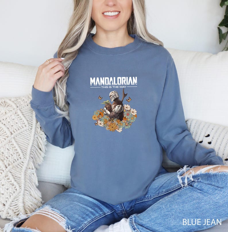 A woman wearing a blue sweatshirt with a graphic of a character from the Star Wars franchise, specifically Mandalorian, sitting on a couch.