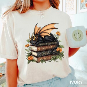 A woman wearing a white t-shirt with a graphic design of a dragon sitting on a stack of books, surrounded by flowers and leaves. She is holding a white mug with a smiley face on it.
