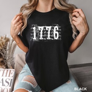 May include: A black short-sleeved t-shirt featuring the white text 1776 printed in a distressed font style. The number 1776 is layered over a background of faint, illegible white text. The shirt has a crew neckline and a standard fit.