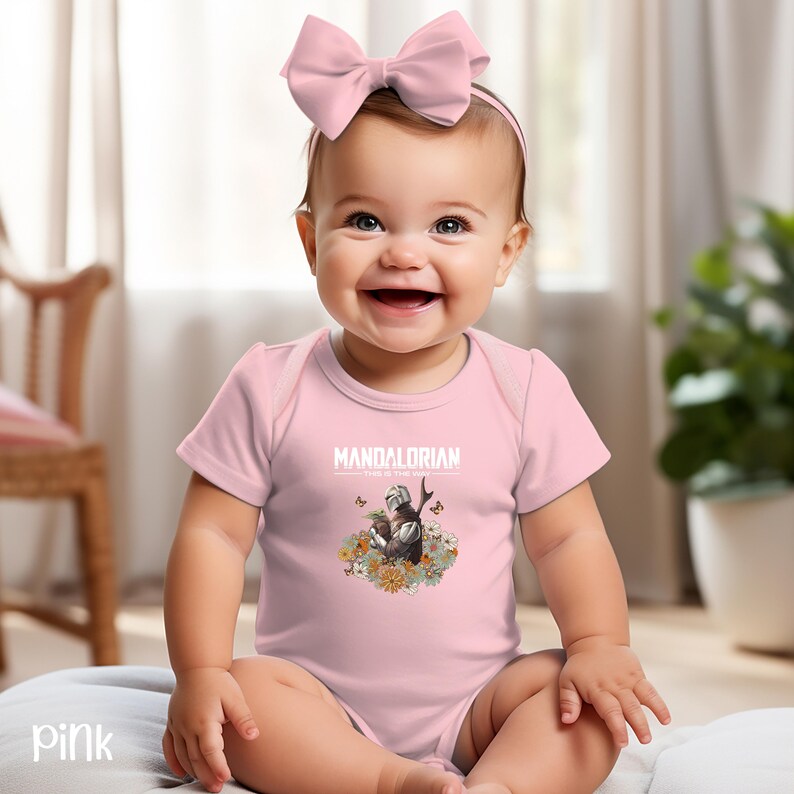 A smiling baby girl wearing a pink outfit with a pink bow and a "MANDALORIAN" t-shirt, sitting on a bed in a room with a potted plant in the background.