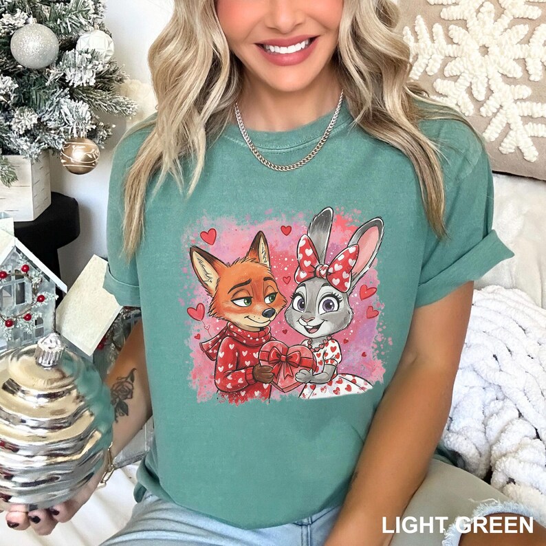 A woman wearing a green t-shirt with a graphic of a fox and a rabbit, both wearing red and white outfits, smiling and holding a small ornament.