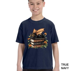 A young boy wearing a navy blue t-shirt with a graphic design of a dragon perched on a stack of books, surrounded by orange flowers.