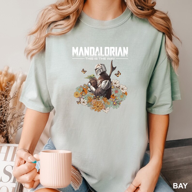 A person wearing a t-shirt with a graphic of a character from the Star Wars series, specifically Mandalorian, holding a cup of coffee.