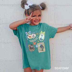 A young girl with pigtails is smiling and making a peace sign with her fingers, wearing a green t-shirt with colorful cartoon characters and designs.