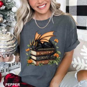 A woman wearing a gray t-shirt with a graphic design of a dragon perched on a stack of books, surrounded by flowers and leaves.